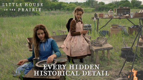 Two girls in prairie dresses sit outdoors beside rustic wooden tables and vintage cooking tools in a grassy field, reenacting a historical pioneer camp scene. A campfire is visible in the foreground, trees in the background.