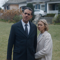 Man and woman standing closely outside on a lawn, both wearing coats, with a large house, stone steps, and a parked car in the background, appearing serious or concerned.