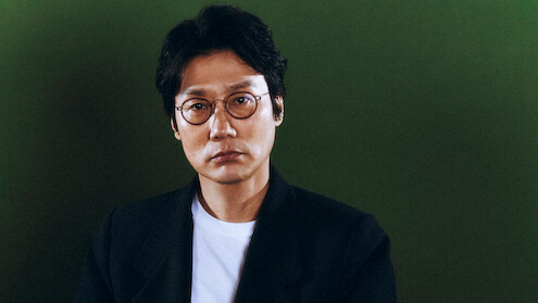 A man with short black hair and round glasses wearing a black blazer and white shirt sits in front of a plain green background, looking directly at the camera with a neutral expression.