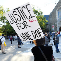 Person holding a large sign reading "Justice for Jane Doe" at an outdoor protest or rally. The event takes place in a plaza in front of a stone building with people gathered and some recording with cameras.