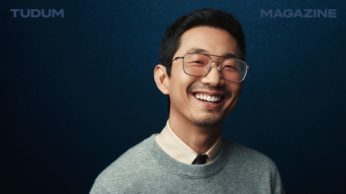 Lee Sung Jin stands in front of a backdrop that shifts from black to dark blue, smiling and looking to camera.
