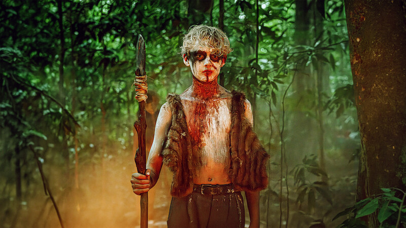 A person with painted face and fur vest stands holding a spear in a dense, misty, green forest, suggesting a primitive or survival setting.