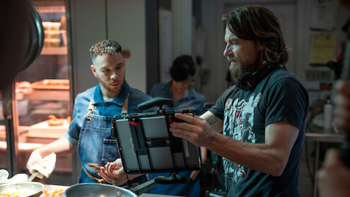 A director on set in casual dress shows an actor a tablet as they work together on a shot.