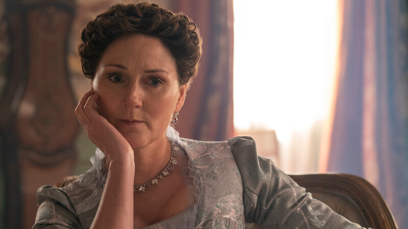 A woman with styled brown hair and vintage clothing sits indoors on a chair, resting her head on her hand, with soft natural light and ornate décor in the background, evoking a historical or period setting.