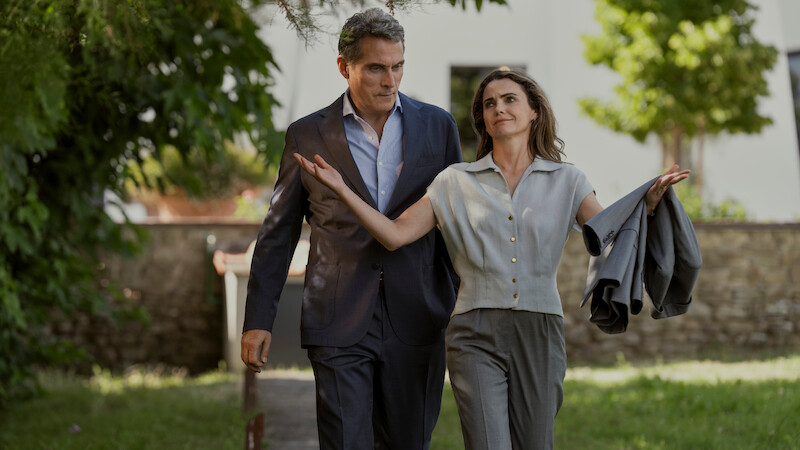 A man in a suit and a woman holding a jacket walk together outside on a sunlit path, surrounded by greenery and trees, with a building in the background.