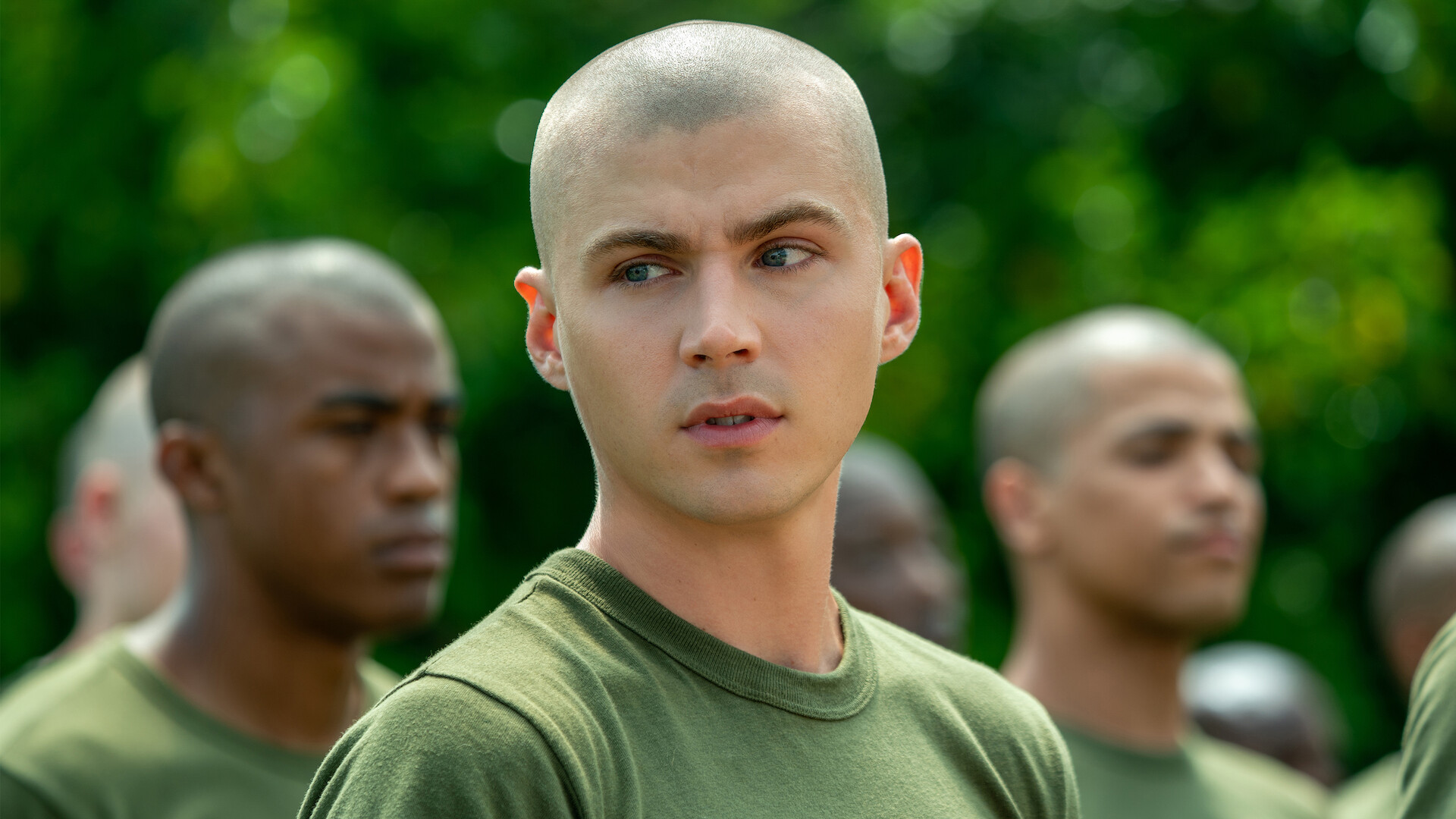 Young man with shaved head in green military shirt stands outdoors among other recruits with similar haircuts, blurred green foliage in background, serious expressions, appearing to be in a training or boot camp setting.