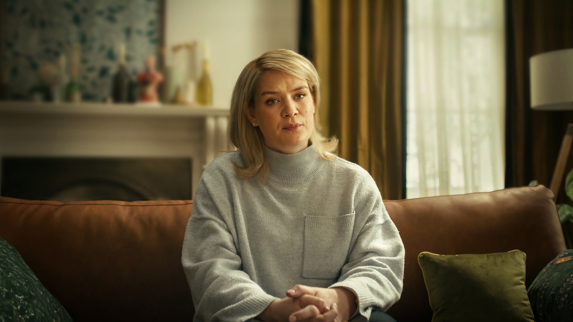 Woman with blond hair sitting on a brown sofa in a cozy living room, hands clasped, wearing a light gray sweater. Background includes a window with curtains, cushions, and a decorated mantelpiece. Natural light fills the room.