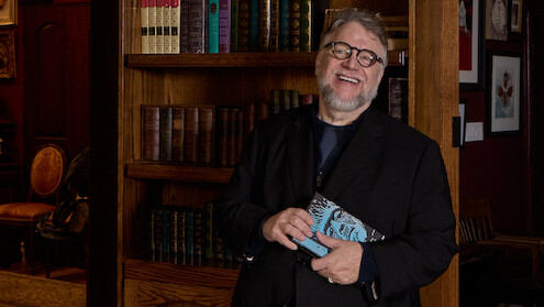 Guillermo del Toro holds a copy of the book of Frankenstein while standing in front of a book shelf.
