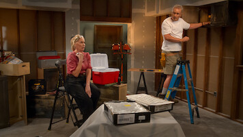 Woman sitting and man standing on ladder in unfinished garage or basement, surrounded by tools, coolers, and boxes, with exposed walls and construction materials, appearing to be in the middle of a home renovation project.