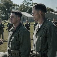 Two Marines stand at attention during a military drill on a grassy field, with several soldiers holding rifles in formation behind them and barracks in the background under a clear sky.