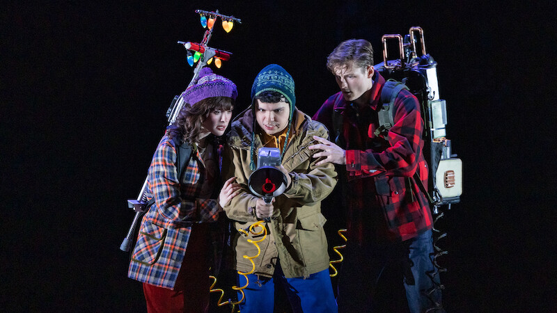 Three young people in winter clothes, holding ghost-busting equipment with glowing lights, stand closely together in a dark, dramatic setting, appearing focused and ready for action.