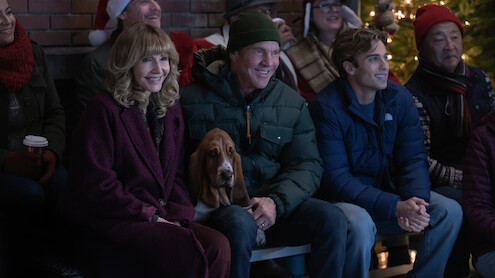 Group of people, including a woman, two men, and a dog, sit together on a bench in a festive indoor setting with a Christmas tree, wearing winter clothes and smiling, enjoying a holiday event.