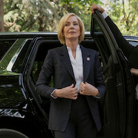 Confident woman in a suit steps out of a black car, adjusting her jacket, with wooded greenery in the background and another person holding the door open.