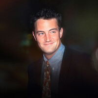 A young man in a suit and patterned tie smiles slightly, standing in a dimly lit environment with soft, colorful bokeh lights in the background, suggesting an evening or nighttime urban setting.