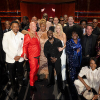 A diverse group of people, including men and women dressed in formal and stylish attire, pose together on a stage in a warmly-lit theater environment, suggesting a celebratory or entertainment industry event.