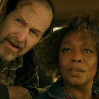 A concerned older man and a woman sit closely together inside a car, both looking intently at something outside, suggesting suspense or curiosity in a dramatic setting.