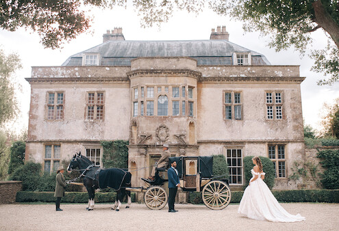Bride in a white gown stands near a vintage horse-drawn carriage outside an elegant, historic mansion. Lush greenery frames the scene, evoking a romantic, fairytale atmosphere. The mood is dreamy and regal.