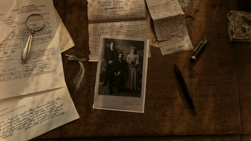 Old sepia-toned family photo on a wooden desk surrounded by vintage papers, handwritten letters, a magnifying glass, pen, pencil, newspaper clippings, and a small ornate box, creating an atmosphere of research or investigation.