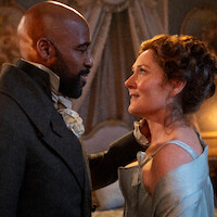 A man and woman in historical costumes gaze at each other affectionately in an ornate room with elegant drapes and soft lighting, suggesting a romantic or intimate moment in a period drama setting.