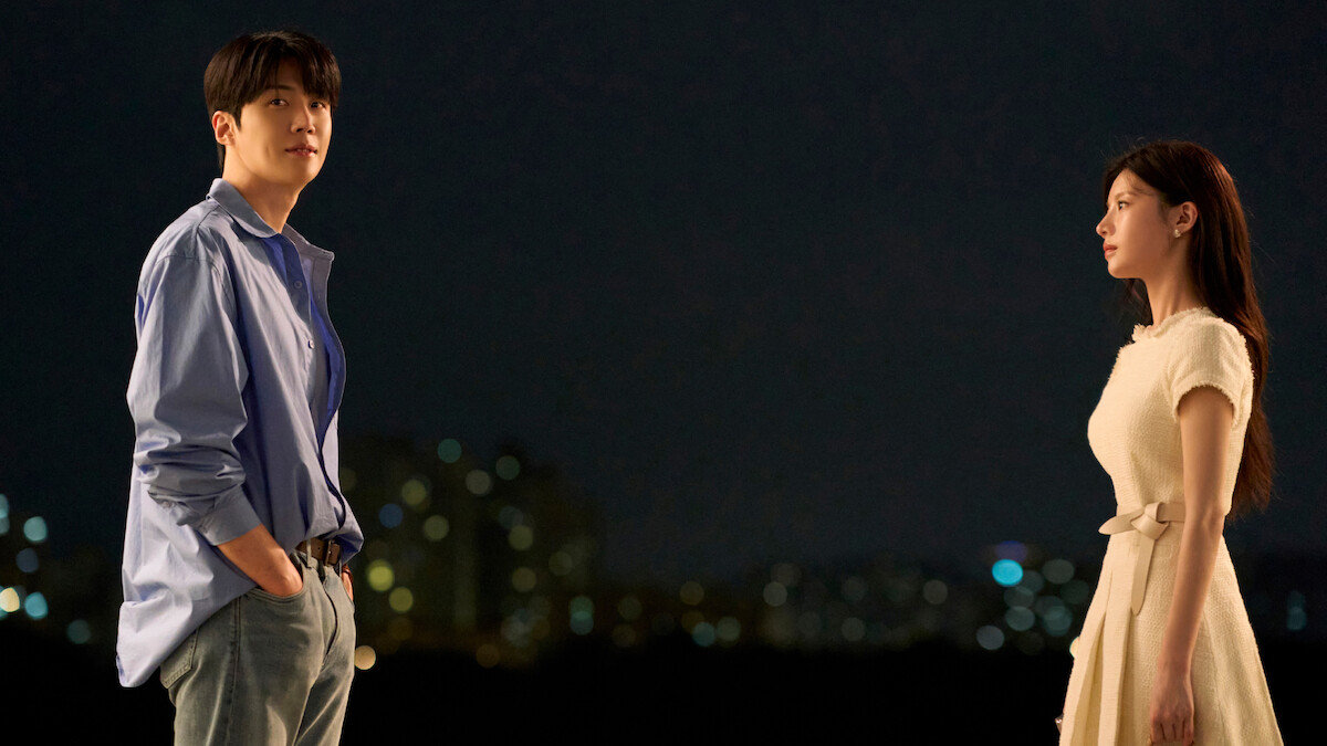 A man and a woman stand facing each other outdoors at night, city lights blurred in the background, creating a dramatic and contemplative atmosphere.