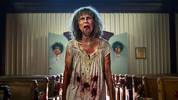 A disheveled, bloodied woman with gray hair stands in the aisle of a decorated church, with pews on either side and wreaths on the doors in the background, looking distressed or possibly zombified.
