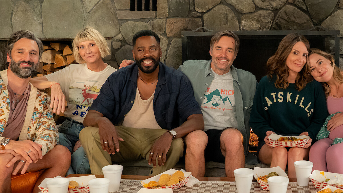 Six adults sitting close together on a couch in a cozy room with a stone fireplace, enjoying a casual meal with trays of food and drinks, smiling and relaxed in a friendly, informal setting.
