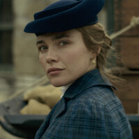 Woman in vintage blue clothing and hat looks over her shoulder outdoors, with a blurred background featuring sacks and bricks, suggesting a historical or period setting.