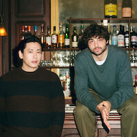 Teo Yoo leans against, and Noah Centineo sit atop the bar looking directly to camera.