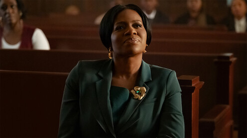 Woman in a green suit sitting on a wooden pew in a church or courtroom, looking upward thoughtfully, with people blurred in the background.
