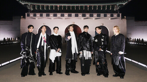 V, JIN, SUGA, RM, JUNG KOOK, J-HOPE, and JIMIN dressed in fashionable black and white outfits stand outdoors at night in front of an ornate, traditional East Asian building with arches and a large decorative roof.