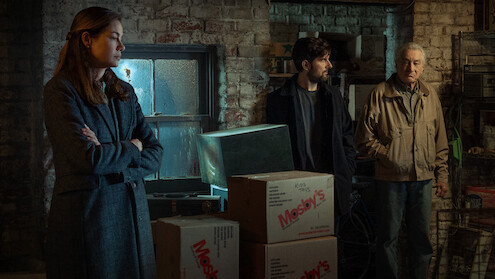 Three people stand in a dimly lit, cluttered basement or storage room with brick walls and stacked boxes. The mood appears tense or serious, and the setting looks old and industrial.