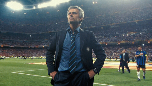 Soccer coach in a suit standing confidently on a stadium field, with players and a large crowd in the background during a nighttime sports event.