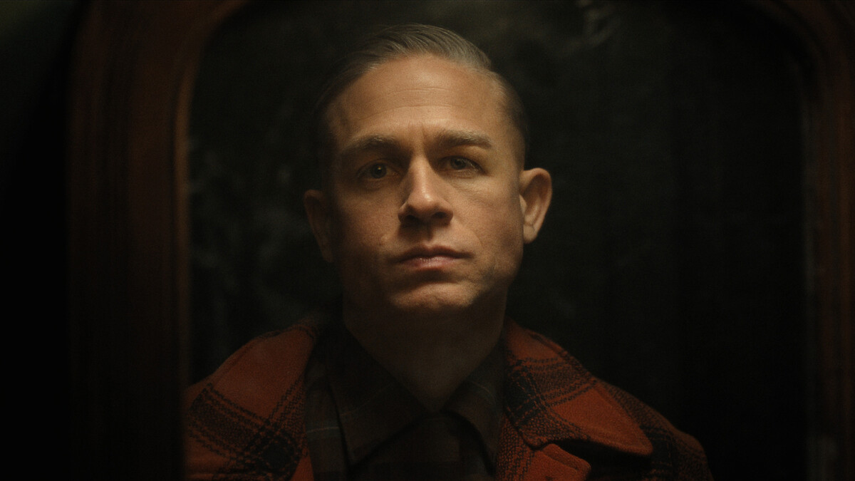 Ed Gein in a plaid jacket looks at his reflection in the mirror in a dark room.