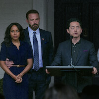 Three adults stand together outdoors; one man gives a speech at a podium, while another man in a suit comforts a concerned woman beside him. The setting appears formal, possibly a press conference or announcement.