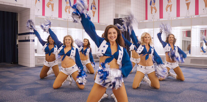 Cheerleaders com uniforme do DCC ensaiam em formação triangular em uma sala de prova e erguem com confiança a mão direita com pompons para o alto.