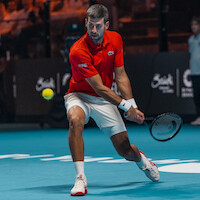 Male tennis player in a red shirt and white shorts hitting a backhand shot on an indoor hard court during a match, with the tennis ball in motion and an audience in the background.