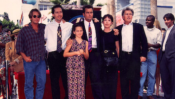A group of people posing together outdoors on a red carpet, dressed in formal and casual attire, with palm trees and buildings in the background during a sunny day event.