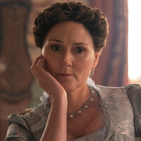 A woman with styled brown hair and vintage clothing sits indoors on a chair, resting her head on her hand, with soft natural light and ornate décor in the background, evoking a historical or period setting.