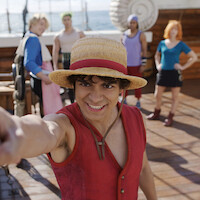Smiling young man in a straw hat and red vest poses on a wooden ship deck with four friends behind him, ocean visible in the background, all in colorful costumes, suggesting an adventurous nautical setting.