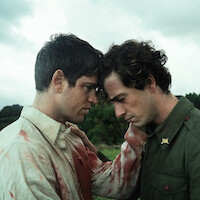 Two injured men in bloodstained uniforms comfort each other, standing close with foreheads touching outdoors under a cloudy sky, conveying emotion and solidarity in a natural, green environment.