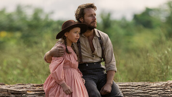 A man and a young girl in old-fashioned clothing sit on a large log outdoors. The man has his arm around the girl, and they both look thoughtfully into the distance, surrounded by greenery and soft natural light.