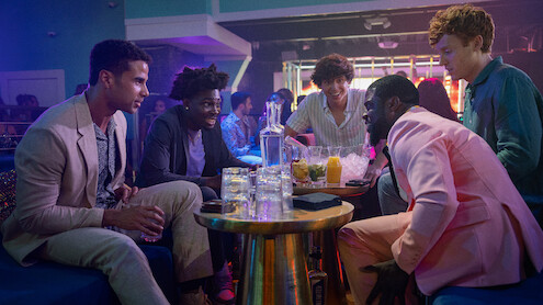 Five men sitting around a table with drinks, laughing and talking in a lively nightclub with colorful lighting and a modern, vibrant atmosphere.