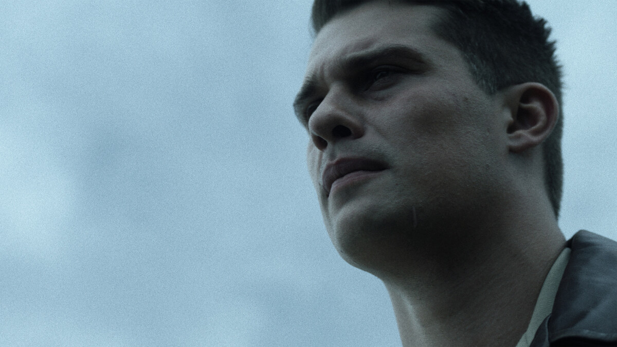 Young man with short dark hair in a serious expression, shown in close-up against a cloudy sky, outdoors in natural light.