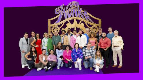 Grupo de pessoas posando juntas em frente a um grande portal decorativo com a palavra “Wonka”, algumas de pé e outras sentadas em um tapete roxo, vestidas com roupas coloridas e sorrindo em um estúdio ou espaço de evento.