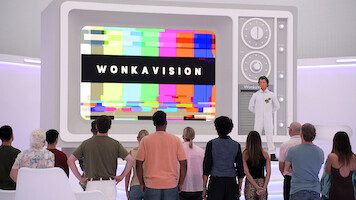 A group of people watch a large retro television screen displaying “WONKAVISION” with colorful test patterns, in a futuristic, white room. A person in a white lab coat stands beside the TV, leading the presentation.