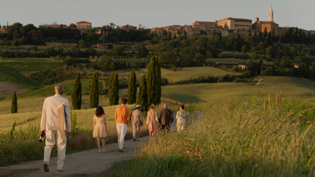 A group of people walk down a country path through green fields at sunset, heading toward a distant hilltop village, evoking a peaceful and warm atmosphere.