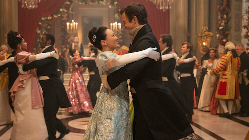 Um casal em elegantes trajes de época dança em um grande salão, cercado por outros casais dançando e convidados em vestes elaboradas, com decorações deslumbrantes, flores e lustres, em uma atmosfera festiva de época.