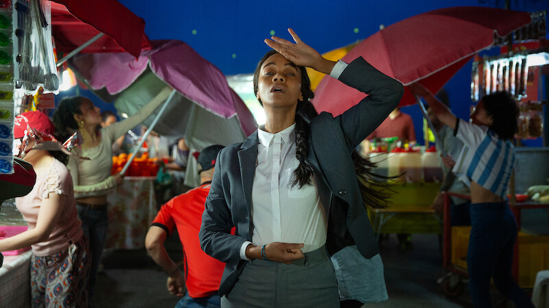 Rita (Zoe Saldaña) wears a white shirt and gray suit and dances at an outdoor market.