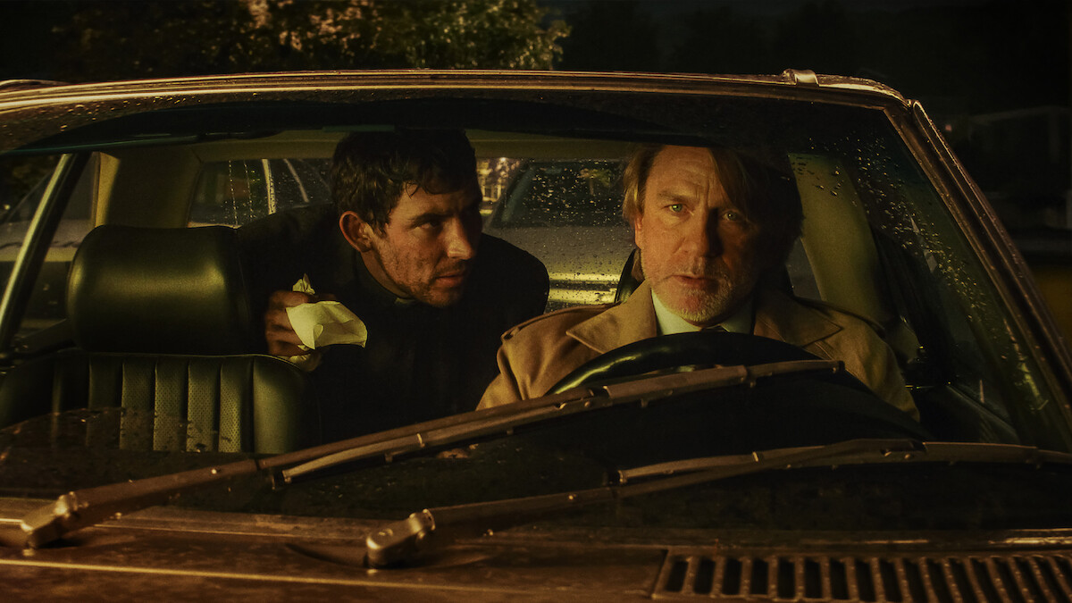 Two men sit in a car at night, illuminated by dim streetlights and rain on the windshield. The driver looks concerned while the passenger leans in with urgency, creating a tense and suspenseful atmosphere.