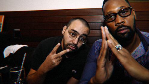 Two men sitting indoors at a table, one making a peace sign and the other with hands pressed together, both wearing glasses in a dimly-lit setting with wooden paneling in the background.